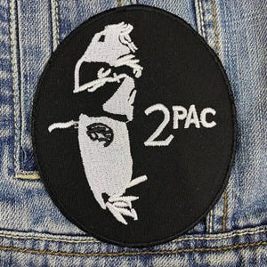 Puede incluir: Parche bordado negro, de forma ovalada, con una silueta blanca de una persona y el texto "2PAC". El parche está sobre un fondo de mezclilla.