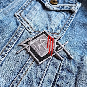 May include: Close-up of a light blue denim jacket with a patch. The patch is a gray and black geometric design with three red vertical lines. The jacket has a pocket and visible stitching.