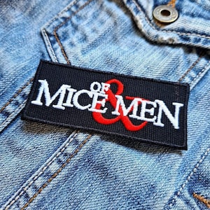 May include: A black rectangular patch featuring the text "MICE OF MEN" in white, with a red ampersand. The patch is attached to a light blue denim jacket, showing stitching and a button.