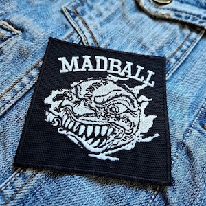 May include: Black square patch with the word "MADBALL" in white block letters above a detailed white illustration of a snarling face. The patch is on a denim jacket.