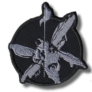 Puede incluir: Parche bordado con un diseño de insecto gris estilizado sobre un fondo circular negro. El insecto tiene alas detalladas y un cuerpo central, con un elemento en forma de bandera en la parte superior. El parche tiene un borde negro.