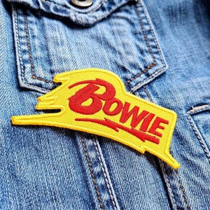 Könnte beinhalten: Ein gelb-roter, gestickter Aufnäher mit dem Namen "Bowie" in einer stilisierten Schriftart. Der Aufnäher ist rechteckig mit einem geschwungenen Design und ist auf einer Jeansjacke befestigt.