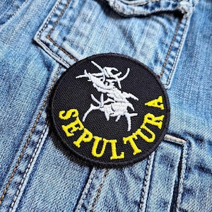 Puede incluir: Un parche redondo negro con la palabra "SEPULTURA" en letras amarillas. El parche presenta un diseño abstracto blanco en el centro. El parche está cosido a una chaqueta vaquera.