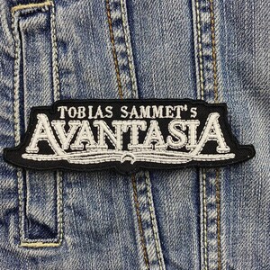 Puede incluir: Parche bordado en blanco y negro con el texto "TOBIAS SAMMET'S AVANTASIA" sobre un fondo de mezclilla. El parche tiene un borde negro y letras blancas, y es rectangular con un borde inferior curvo.