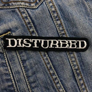 May include: A black embroidered patch featuring the word "DISTURBED" in white, bold lettering. The patch is rectangular and is set against a blue denim background, with visible stitching.