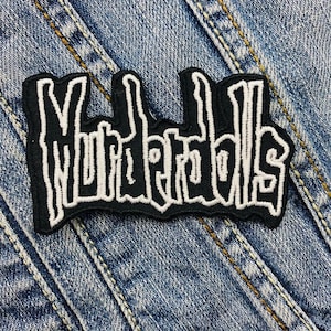 May include: A black and white embroidered patch with the text "Murderdolls" in a stylized font. The patch is rectangular with a black border and is placed on a denim background.