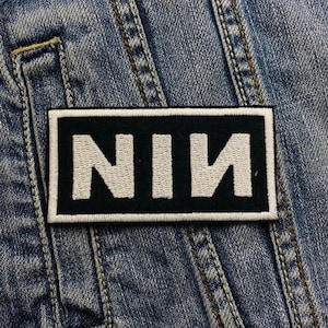 May include: A rectangular black patch with the letters "NIN" embroidered in white. The patch has a white border and is sewn onto a denim jacket. The denim jacket is blue with visible stitching.
