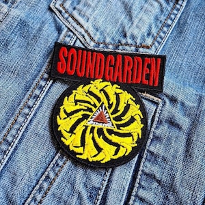 Op de afbeelding: Een spijkerjasje met twee geborduurde patches. De bovenste patch leest "SOUNDGARDEN" in rode letters op een zwarte achtergrond. De onderste patch is een geel en zwart cirkelvormig ontwerp met een bruine driehoek in het midden.