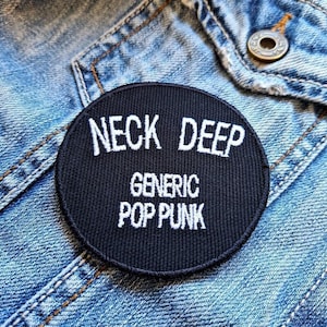 Puede incluir: Un parche redondo negro con texto blanco bordado. El texto dice "NECK DEEP" encima de "GENERIC POP PUNK". El parche está colocado sobre una chaqueta vaquera azul, con costuras visibles y un botón metálico.