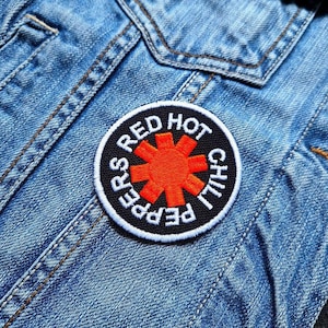 Può includere: Una toppa rotonda con la scritta "RED HOT CHILI PEPPERS" in bianco su sfondo nero e un disegno a forma di asterisco arancione. La toppa è cucita su una giacca di jeans blu.