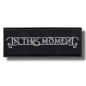 Pode incluir: Patch retangular preto com o texto bordado "IN THIS MOMENT" em branco, com letras estilizadas. O patch tem uma borda preta e um efeito de sombra.