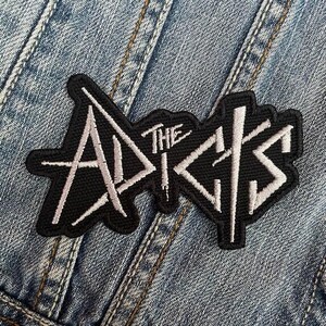 May include: A black embroidered patch featuring the words "THE ADDICTS" in white, distinctive lettering. The patch is attached to a denim jacket, showing the fabric's texture and stitching, with a light blue hue.