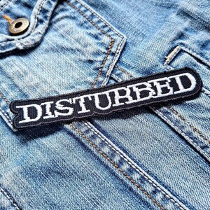 May include: A black and white embroidered patch with the word "DISTURBED" in white block letters. The patch is rectangular and has a black border. It is placed on a light blue denim jacket.