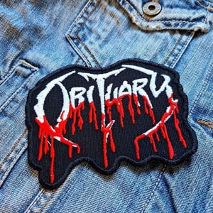Könnte beinhalten: Ein schwarzer, gestickter Aufnäher mit dem Wort "Obituary" in Weiß und tropfenden roten Akzenten. Der Aufnäher ist auf eine Jeansjacke genäht. Der Aufnäher hat einen schwarzen Rand.
