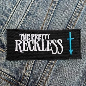 May include: A black rectangular patch featuring the white text "THE PRETTY RECKLESS" and a turquoise cross with a downward arrow. The patch is set against a denim background.