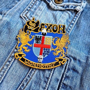 May include: Embroidered patch with the word "SAXON" in gold lettering. The patch features a blue shield with a red cross and gold heraldic eagles. The bottom of the patch reads "LIONHEART" in gold lettering, with a skull in the center.