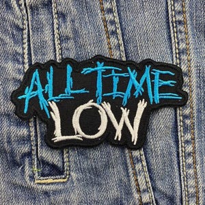May include: A black embroidered patch with the words "ALL TIME LOW" in a stylized font. The text "ALL TIME" is in light blue, and "LOW" is in white. The patch is sewn onto a denim jacket.