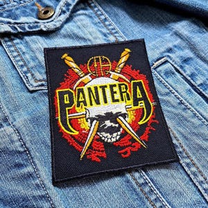 May include: Black rectangular patch with the word "PANTERA" in yellow and red, featuring a skull and crossed swords design. The patch is on a denim jacket.