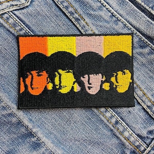Può includere: Toppa ricamata raffigurante quattro volti con capelli neri su uno sfondo di strisce verticali arancioni, gialle, rosa e gialle. La toppa rettangolare è su una giacca di jeans. La toppa è un design vibrante e colorato.