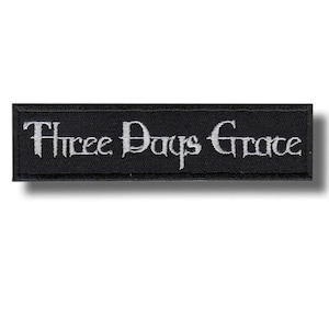 May include: Black rectangular patch with the text "Three Days Grace" embroidered in white. The text is in a stylized font. The patch has a black border and is likely an accessory for clothing or bags.