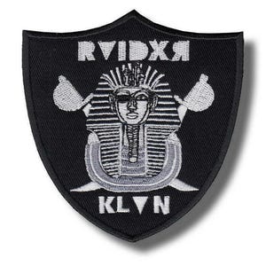 Peut inclure: Écusson noir en forme de bouclier avec texte et motif brodés en blanc. Le motif représente une tête de pharaon, des outils croisés et les textes "RVIDXR" et "KLVN". L'écusson est probablement destiné aux vêtements ou accessoires.