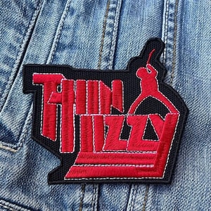 May include: Embroidered patch featuring the band name "Thin Lizzy" in red, outlined in white, on a black background. The patch includes a red silhouette of a guitar player. The patch is sewn onto a denim jacket.
