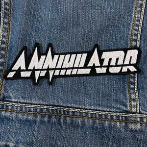 May include: A black and white embroidered patch with the word "Annihilator" in a stylized font. The patch is rectangular and has a jagged design element. The patch is sewn onto a blue denim jacket.