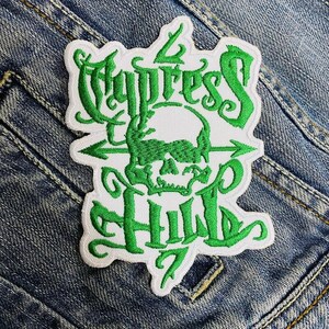 May include: Embroidered patch with the words "Cypress Hill" in green script, featuring a green skull and crossbones design. The patch has a white background and is set against a denim background.