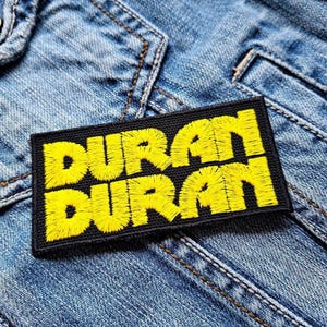 Op de afbeelding: Een rechthoekige zwarte patch met de geborduurde tekst "DURAN DURAN" in helder geel. De patch is genaaid op een lichtblauwe spijkerjas, met zichtbare stiksels en stoftextuur.