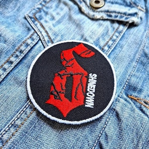 Pode incluir: Remendo redondo preto com um gráfico vermelho e a palavra SHINEDOWN em branco. O remendo tem uma borda branca e está em uma jaqueta jeans.