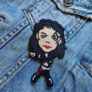 May include: Embroidered patch featuring a cartoon depiction of a person in a black outfit with a red stripe, black hair, and red lips. The patch is sewn onto a blue denim jacket.