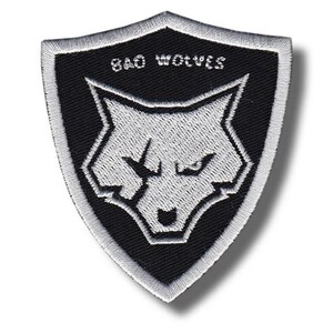 May include: A black and white embroidered patch in a shield shape. The patch features a wolf head design and the text "BAD WOLVES" in white. The patch has a white border and is likely for clothing or accessories.