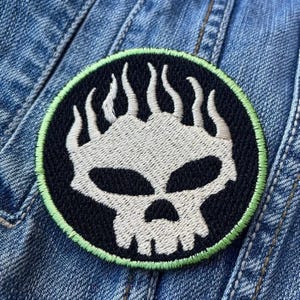 May include: Embroidered patch with a skull and flame design on a black background. The skull is white, and the flames are stylized. The patch has a green border and is sewn onto denim fabric.