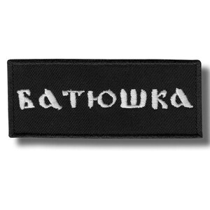 May include: A black rectangular patch with white embroidered text that reads "БАТЮШКА". The patch has a textured surface and a stitched border. The text is in a stylized font.