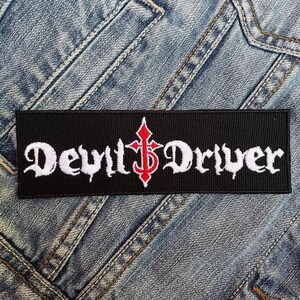 Puede incluir: Un parche rectangular negro con las palabras "DevilDriver" bordadas en letras góticas blancas. Una cruz roja con un signo de dólar está entre las palabras. El parche está sobre un fondo de mezclilla.