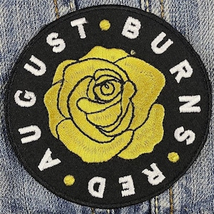 以下が含まれることがあります： 黒色の刺繍入り円形パッチ。白い文字で「AUGUST BURNS RED」と書かれ、黄色のバラを囲んでいます。バラは黒いアウトラインで詳細に描かれています。パッチはデニム生地に縫い付けられています。大胆なデザインです。