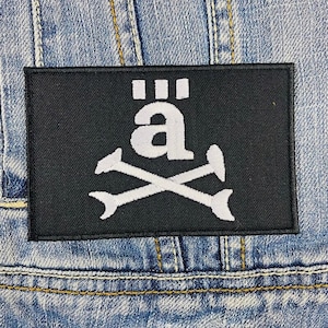 May include: Black rectangular patch with a white design featuring the letter "ä" above crossed bones. The patch is sewn onto a light blue denim jacket, with visible stitching and seams.