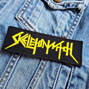 Puede incluir: Un parche rectangular negro con el texto amarillo "SKELETONWITCH" está cosido en una chaqueta vaquera. El parche tiene un aspecto texturizado y el texto está en una fuente estilizada y angular.