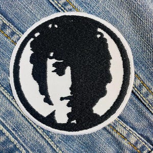 May include: A round, embroidered patch featuring a black silhouette of a person's profile against a white background. The patch is bordered by a white edge and is set against a blue denim background.