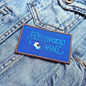 Könnte beinhalten: Ein rechteckiger, blau bestickter Aufnäher mit den Worten "Fleetwood Mac" in hellblauer Schrift und einer kleinen Grafik. Der Aufnäher hat einen braunen Rand und ist auf einer Jeansjacke angebracht.