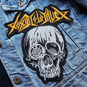 Pode incluir: Patch bordado com um design de caveira detalhado, com um olho e dentes estilizados. O patch tem uma borda preta e um logotipo amarelo e preto na parte superior. O patch é costurado em uma jaqueta jeans.
