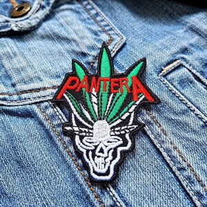 Può includere: Toppa ricamata con la scritta "PANTERA" in rosso, foglie verdi e un teschio. La toppa è in bianco e nero ed è cucita su una giacca di jeans.