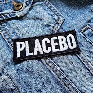 May include: A black rectangular patch with the word "PLACEBO" embroidered in white, affixed to a light blue denim jacket. The jacket features visible stitching and a button detail.