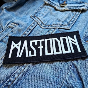 May include: A black rectangular patch with the white word "MASTODON" embroidered on it. The patch is placed on a blue denim jacket. The patch is a music-themed accessory.
