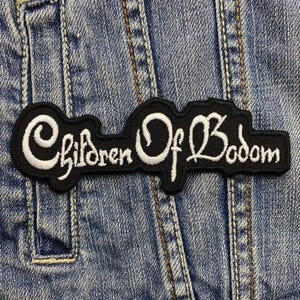 May include: A black patch featuring the text "Children Of Bodom" in white script. The patch is sewn onto a blue denim jacket. The patch has a curved shape.