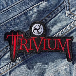 May include: A black patch with the word "TRIVIUM" in red, stylized lettering. Above the word is a white and black circular design. The patch is on a blue denim background.