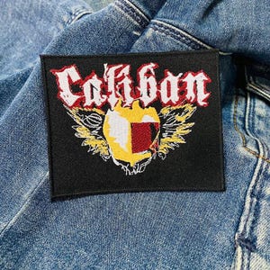May include: A black rectangular patch with the word "Caliban" in red and white gothic lettering. The patch features a yellow and red emblem with wings and is sewn onto a blue denim jacket.