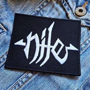 May include: Black rectangular patch with white stylized text and arrow-like designs on a denim jacket. The patch features an embroidered design with a gothic or stylized font. The patch is likely an accessory for clothing or bags.