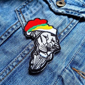 May include: Embroidered patch in the shape of Africa, featuring a lion's head. The patch has a red, green, and yellow top, with black and white detailing. It is sewn onto a blue denim jacket.