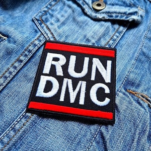 May include: A square black patch with the text "RUN DMC" embroidered in white, framed by a red border. The patch is attached to a light blue denim jacket, representing a classic hip-hop style.